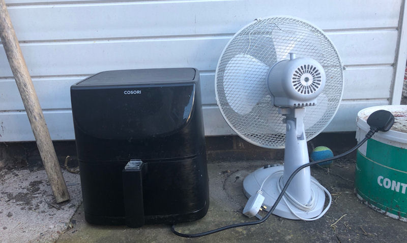 Old, thrown out air fryer sitting next to fan on ground outdoors