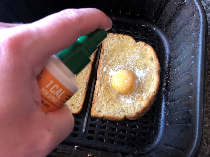 Spraying oil over raw egg sitting on bread slice in air fryer basket