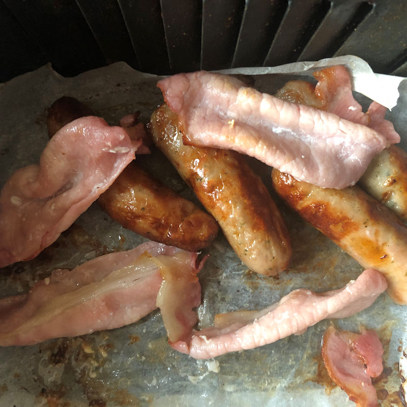 Back bacon piled on top of sausages in air fryer basket