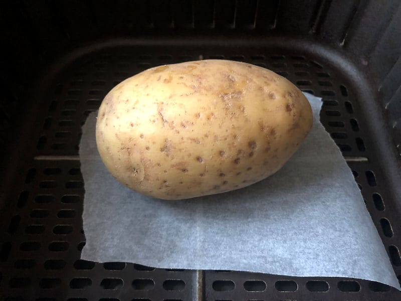 Large single raw potato positioned on parchment paper inside air fryer